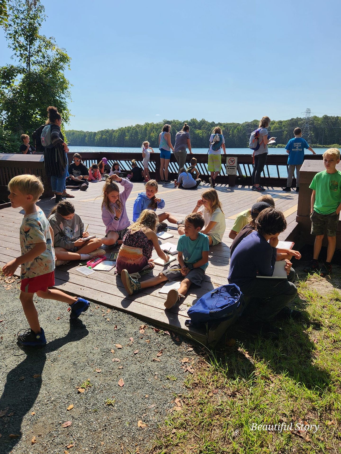 Children and adults enjoying a sunny day on a wooden deck by a lake, surrounded by greenery.