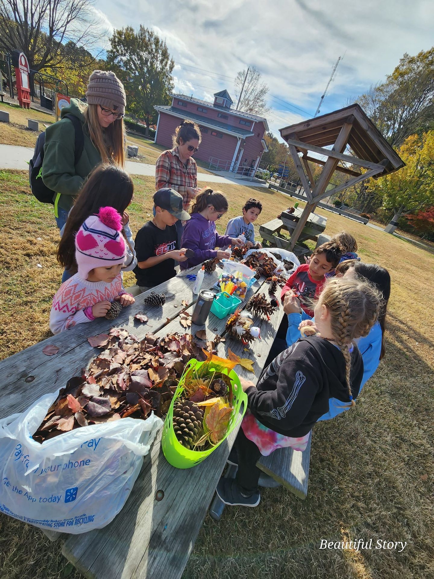 Children and adults crafting with leaves and pinecones at an outdoor table in a park.