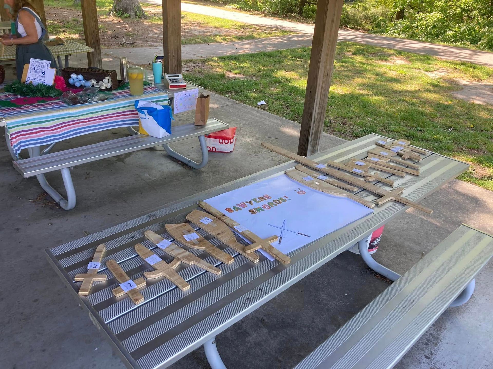 Picnic tables with a display of wooden swords and craft items in a park setting.